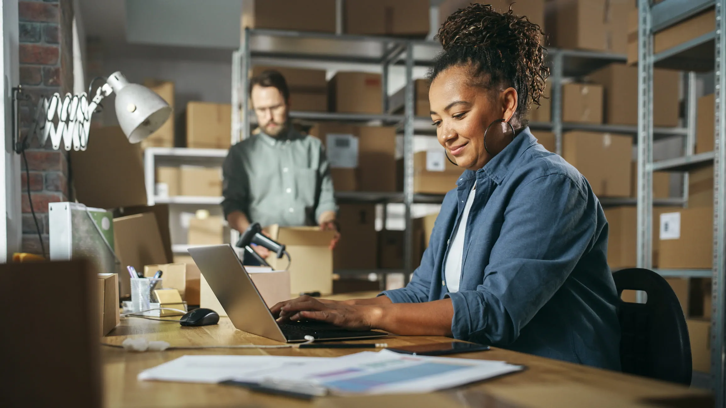 A person sits at a cluttered desk at a laptop in a shipping warehouse. A worker behind them looks at an opened box.
