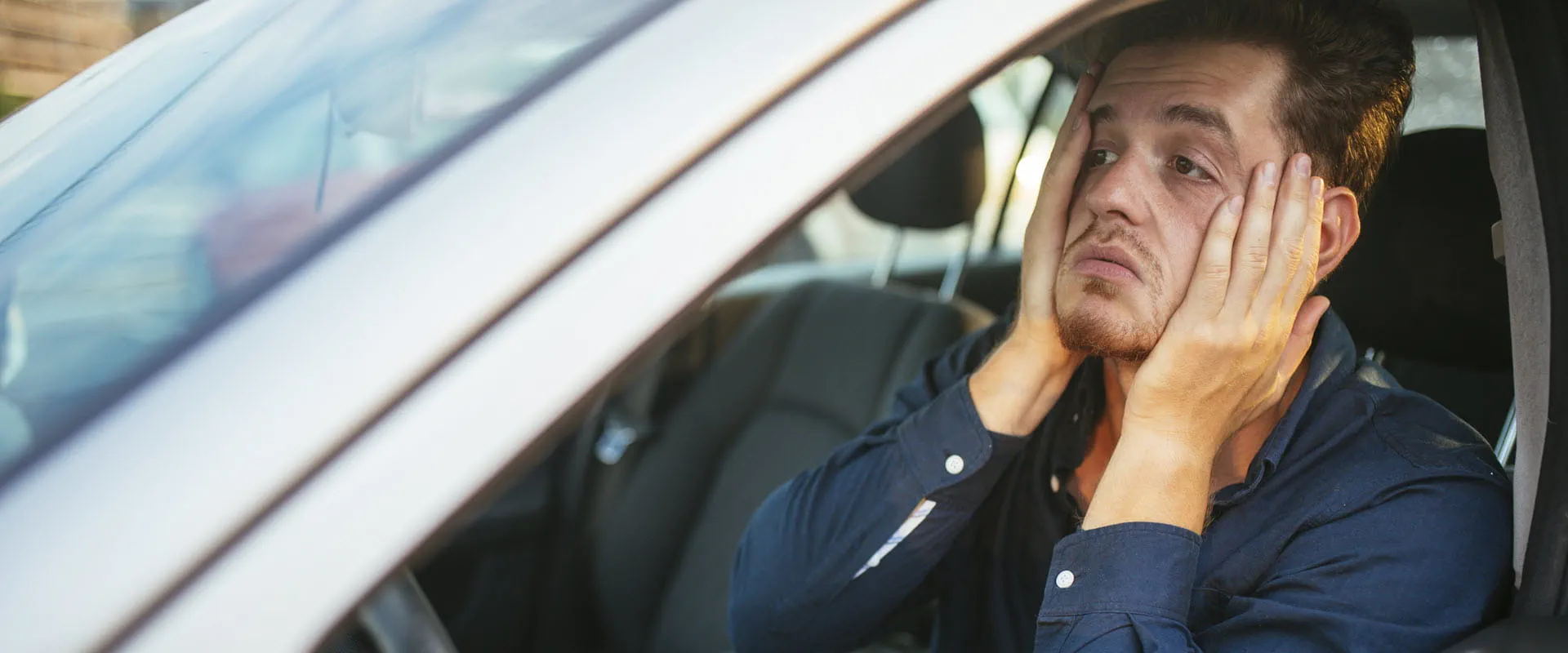 An individual sits in their car, with their hands on their face, seeming overwhelmed.