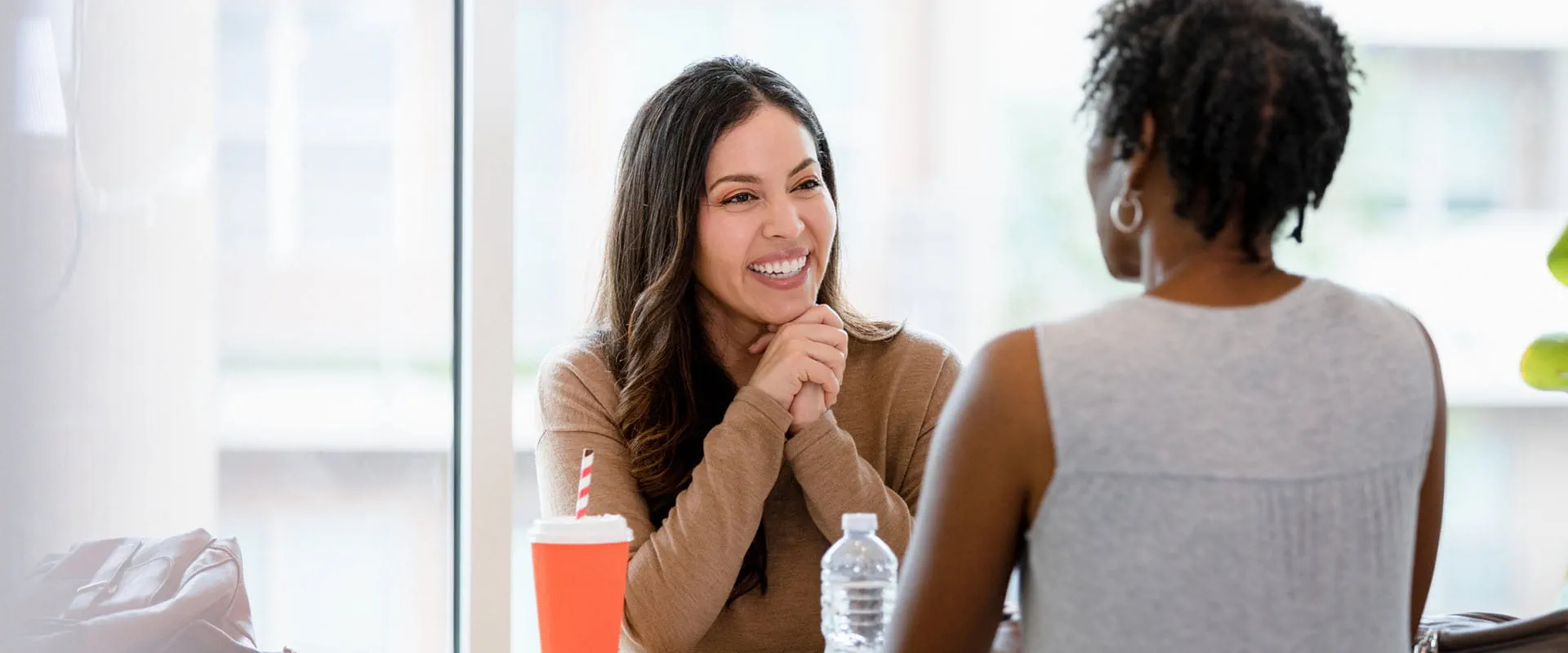 A smiling manager and employee sit across the table from one another in an office building. The table is covered in papers, coffee cup and water bottle.