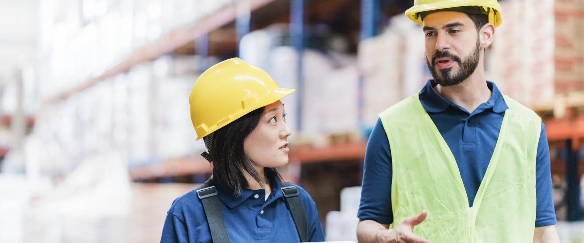 Two workers in hardhats talk, while walking in a warehouse
