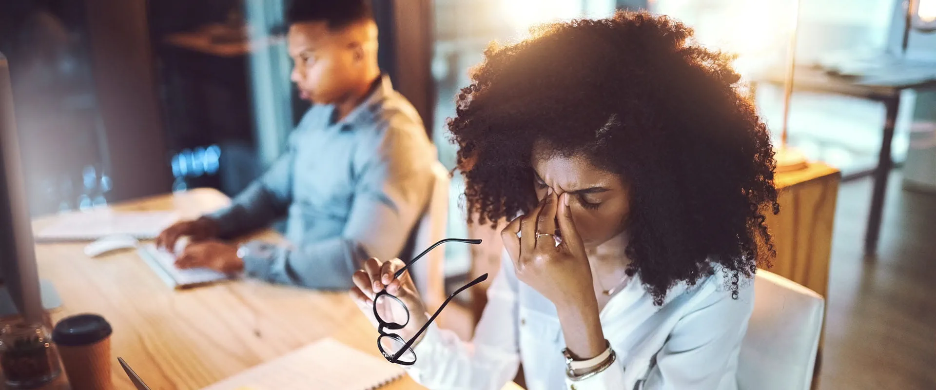 Two workers sit at a table in an office. One is typing at a keyboard. The other has their eyes closed and is rubbing the bridge of their nose.