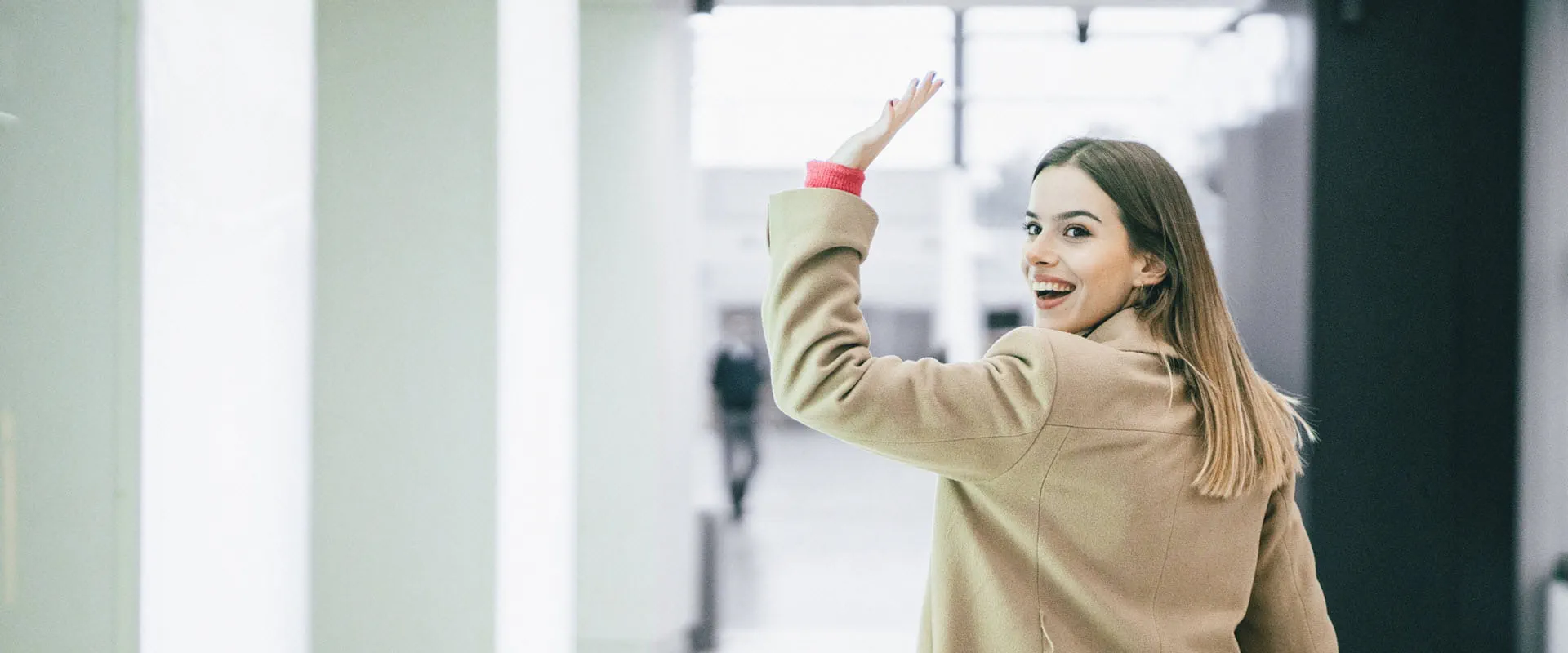 A young woman wearing a beige coat walks down a modern, well-lit hallway while waving and smiling back at the camera.