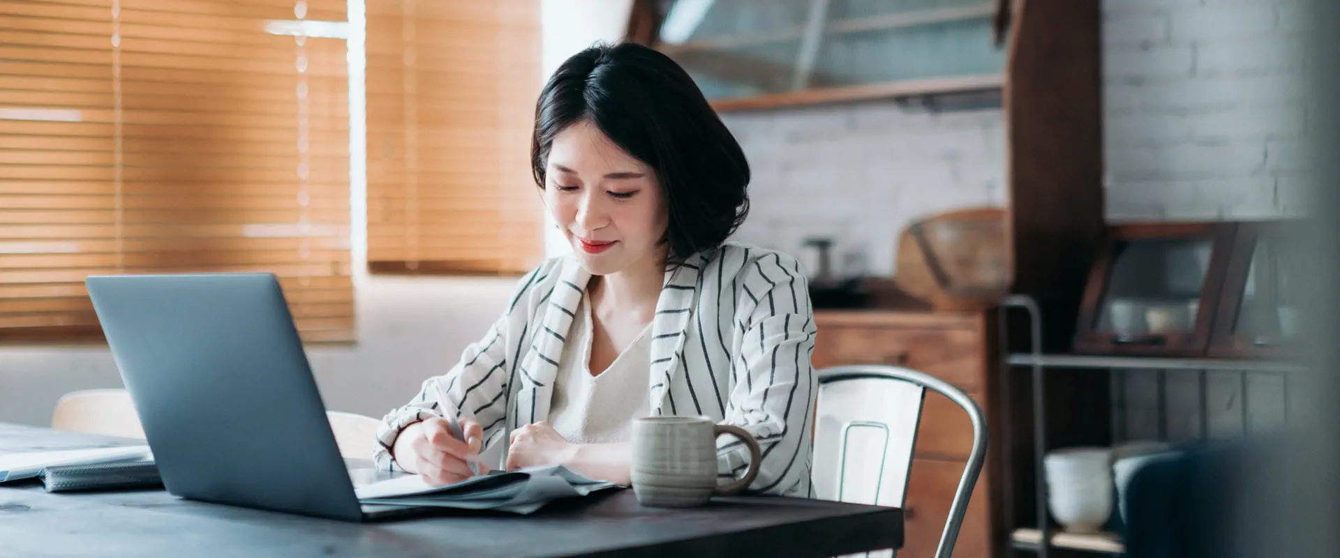 A worker sits at their kitchen table with their laptop, writing something down in a notebook.