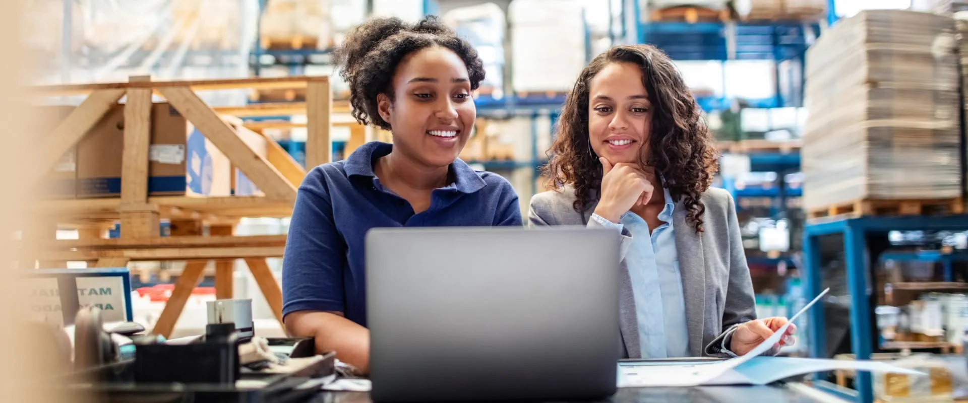 Two female workers in a warehouse, one dress in business attire and the other dressed as a floor worker, look at a laptop together. They are smiling