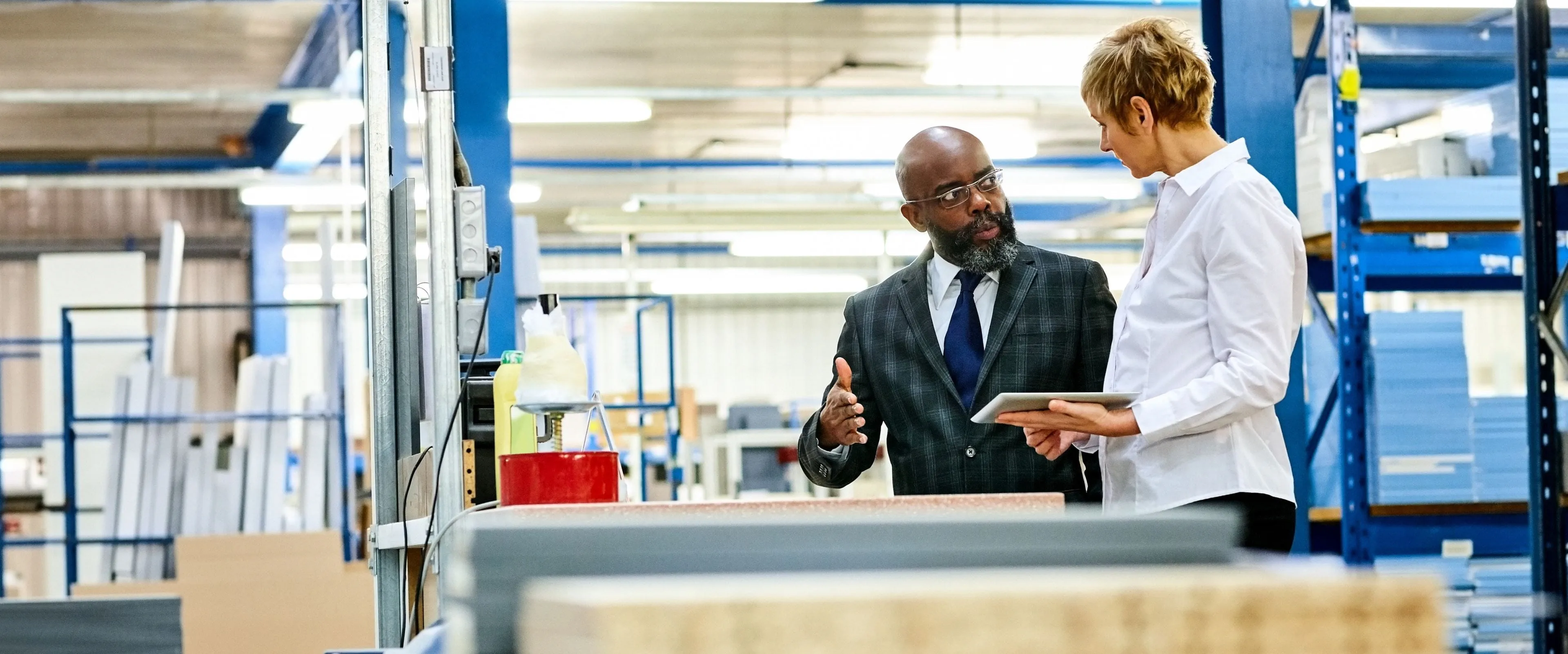 A warehouse floor manager, holding a tablet, speaks with someone in a suit.