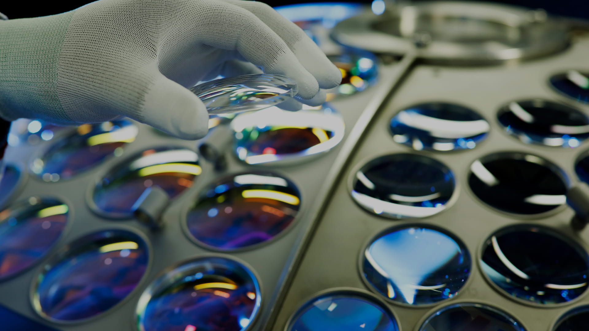 Close-up of a gloved hand handling optical lenses in a precision manufacturing or laboratory setting.