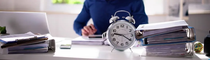 Une employeur travaillant à son bureau, sur lequel est posé un réveil : symbole du  temps à ne pas perdre, pour lancer un recrutement au bon moment.