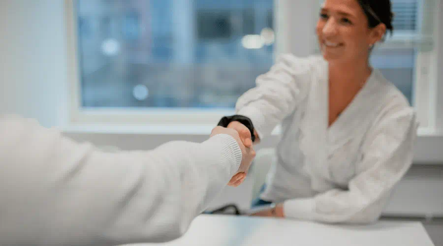 Kvinne smiler mens hun tar håndtrykk med en person under et møte på et kontor.