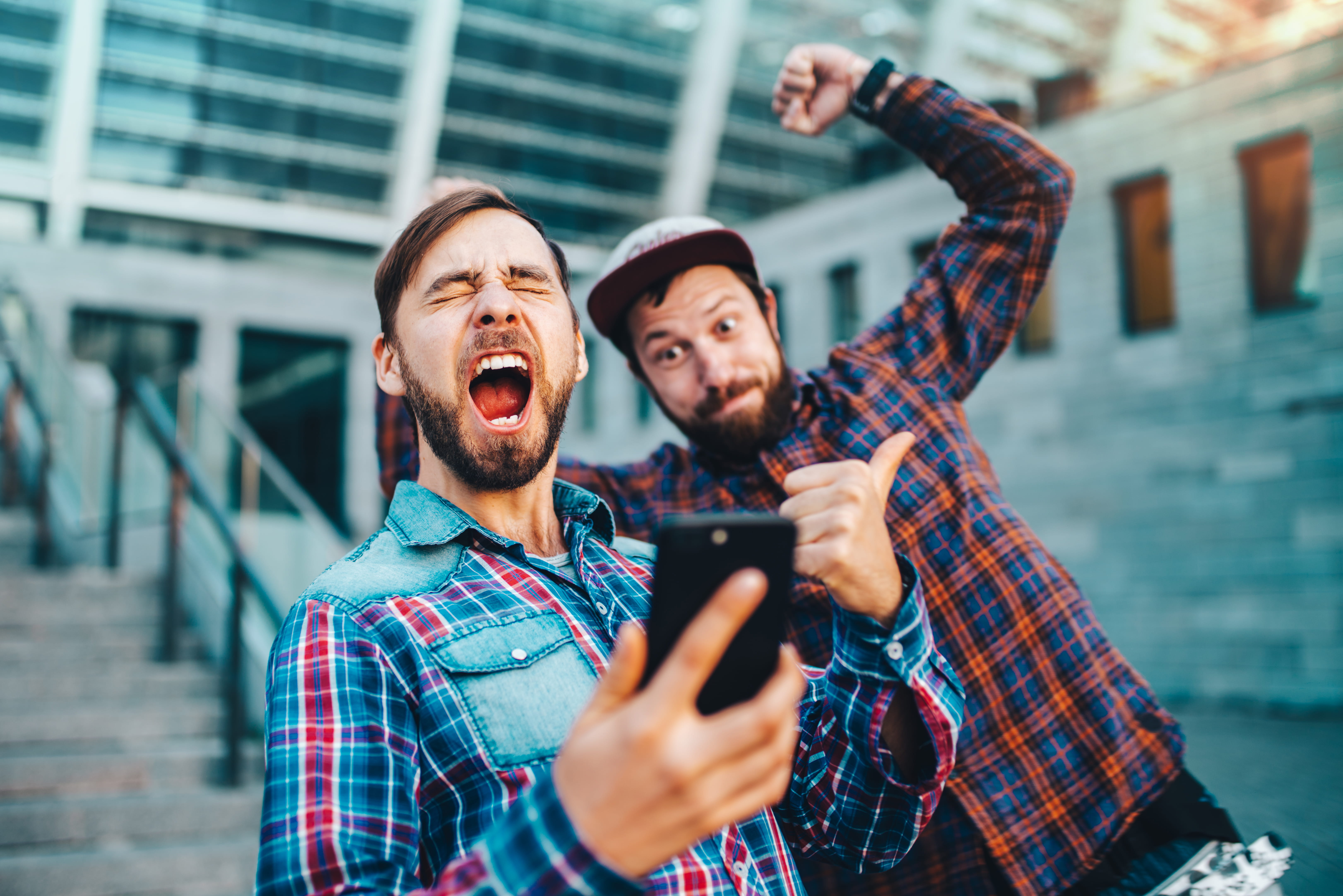 To unge menn i rutete skjorter jubler av glede mens de ser på en mobiltelefon, med en moderne bybakgrunn. To unge menn i rutete skjorter jubler av glede mens de ser på en mobiltelefon, med en moderne bybakgrunn.
