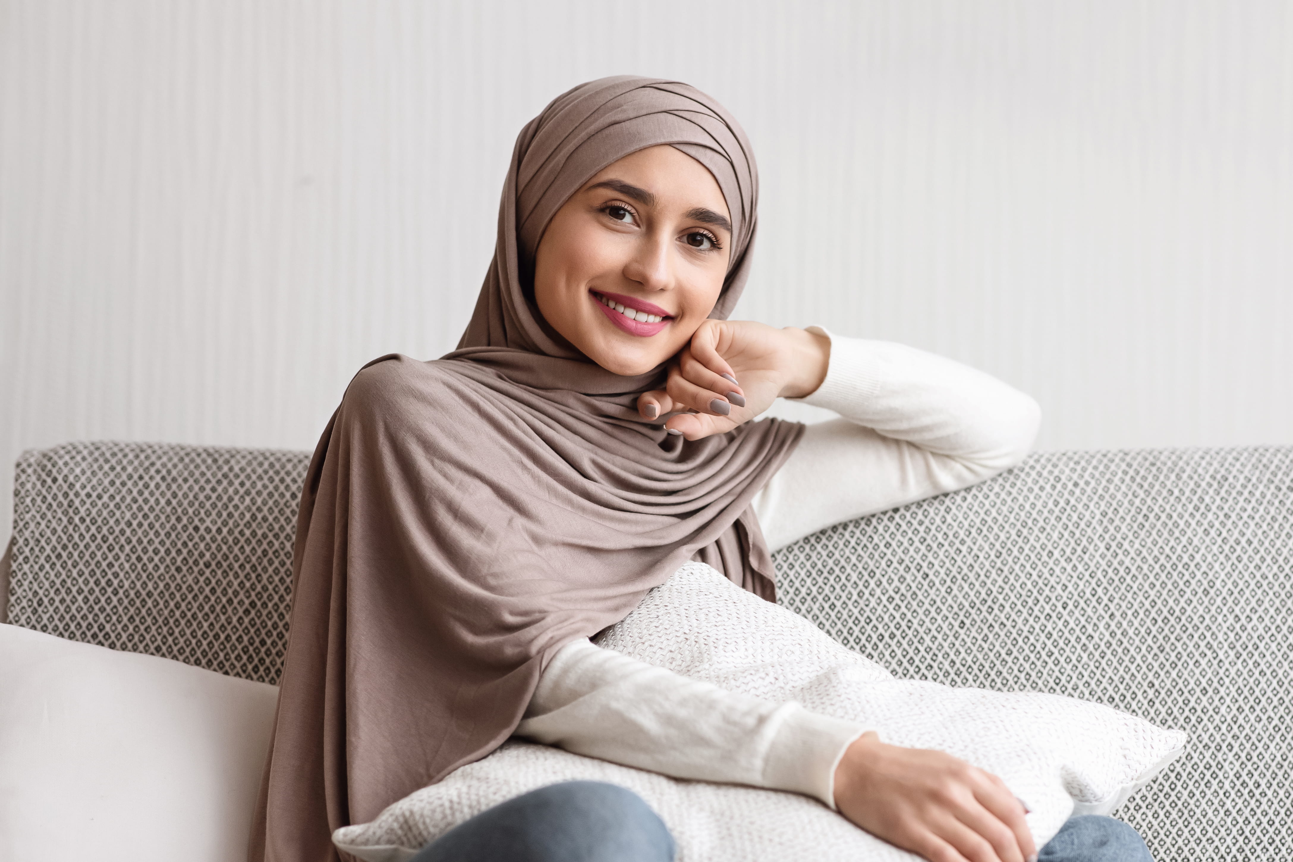 Smilende kvinne i hijab som sitter  komfortabelt på en sofa, kledd i  nøytrale toner. Koselig og  avslappet atmosfære med en myk,  mønstret pute i bakgrunnen.