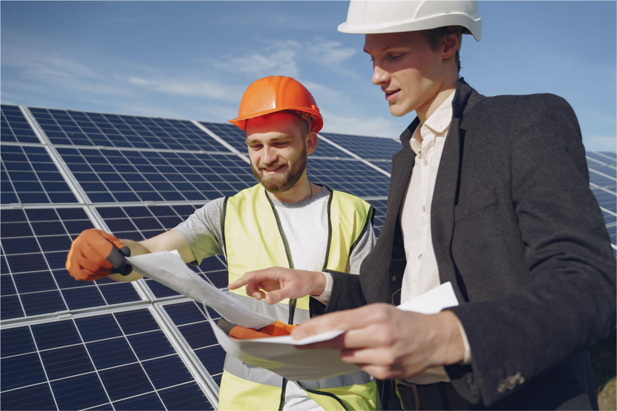 Byggearbeider og prosjektleder inspiserer  tegninger foran et stort solcelleanlegg.  Den ene har på seg hjelm og vernevest,  mens den andre er i dress og hjelm.  Fornybar energi, bærekraft og  byggeprosjekt.